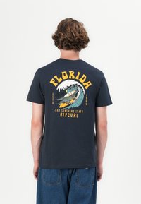 Man wearing black t-shirt with colorful Florida surfing graphic and text on the back, paired with blue jeans, standing against a white background.