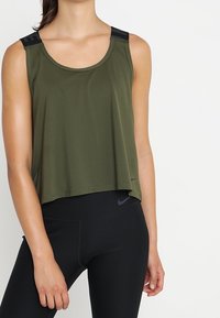 Top de entrenamiento verde aceituna con un corte relajado, acentos en negro en los hombros y un escote redondeado, combinado con leggings negros.