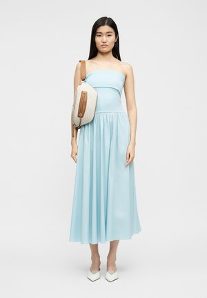 Γυναίκα που φοράει αμάνικο μεσάτο φόρεμα σε ανοιχτό μπλε χρώμα, με λευκή και καφέ τσάντα ώμου και λευκές μυτερές γόβες, στέκεται μπροστά σε ένα απλό φόντο.