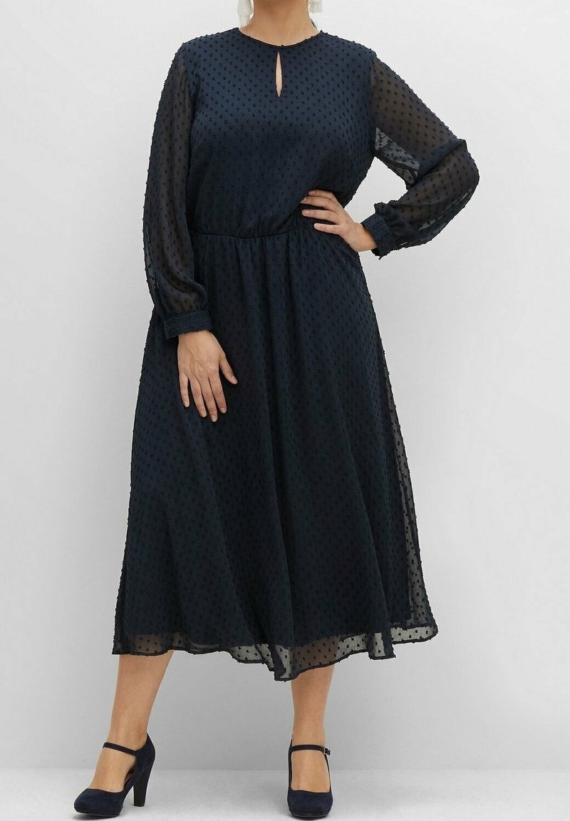 Robe bleu marine à manches longues avec un décolleté en trou de serrure, un motif à pois, une silhouette fluide et des manches en voilage texturé. Portée avec des talons noirs.