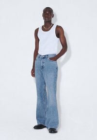 Jeans de mezclilla azul claro acampanados con una cintura alta, bolsillos frontales y un ligero desgastado. Llevados con una camiseta sin mangas blanca y zapatos negros.