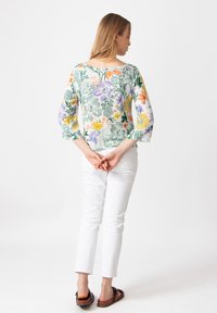 Bloemmotief top met groene, paarse en gele bloemen op een witte achtergrond, voorzien van driekwart mouwen en een brede halslijn. Gepaarde met witte broek.