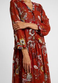 Robe florale rouge à manches longues avec des boutons, coupe ample et détail de taille froncée, portée par une personne croisant les bras sur la poitrine.