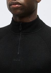 Κοντινό πλάνο ενός ατόμου που φοράει μαύρο πλεκτό πουλόβερ με φερμουάρ στο ένα τέταρτο και διακριτικό λογότυπο "BOSS" στο στήθος.