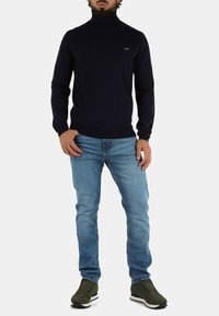 Maglione a collo alto blu marina in maglia ribattuta, abbinato a jeans slim-fit blu chiaro e sneakers verde oliva. Dettaglio logo semplice sul maglione.