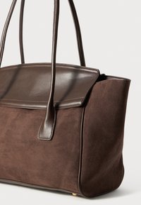 Sac fourre-tout en daim marron avec un rabat en cuir lisse, forme structurée, doubles poignées et détails de couture subtils.