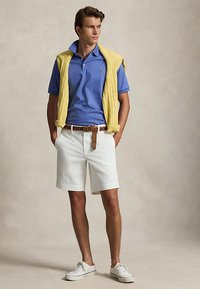 Jeune homme debout portant un polo bleu, un short blanc avec une ceinture tressée marron, un pull jaune drapé sur les épaules et des baskets blanches.