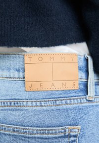 Light blue denim jeans with a tan leather tag featuring embossed text "TOMMY" and "JEANS," stitched at the waistband on the back.