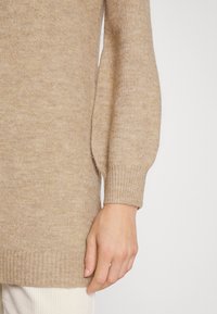 Maglione beige lavorato a maglia con scollatura rotonda, maniche a palloncino e polsini a coste, esposto con una mano su uno sfondo chiaro.
