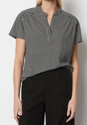 Vrouw draagt een zwart-wit kortermige blouse met patroon en gespleten halslijn, ingestopt in een hoge zwarte broek tegen een effen achtergrond.