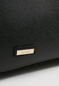 Surface en cuir texturé noir avec une petite étiquette métallique rectangulaire dorée gravée du nom de la marque ALDO.