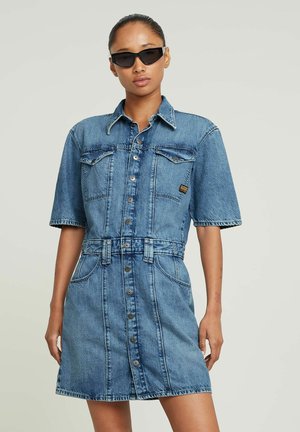 Femme portant une robe en jean à manches courtes avec des boutons sur le devant, des poches poitrine, des passants pour ceinture et des lunettes de soleil noires en forme d'œil de chat, se tenant devant un fond uni.