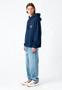 Felpa della marina con ricamo a cuore rosso, tasca a marsupio e cappuccio con cordino, abbinata a jeans blu chiaro e sneakers bianche con dettagli rossi.