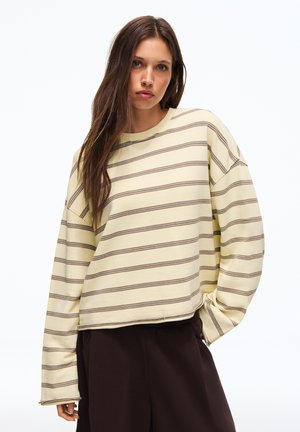 Jonge vrouw met lang bruin haar die een crèmekleurig shirt met lange mouwen draagt met dunne horizontale strepen en donkerbruine broek tegen een witte achtergrond.