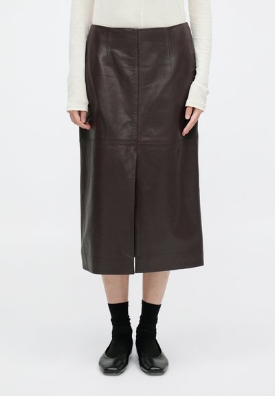 Falda midi de cuero marrón con una abertura frontal, que presenta paneles cosidos y una textura suave. Combinada con zapatos planos negros y calcetas hasta la rodilla.