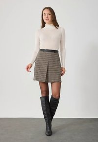 Mini-jupe marron à carreaux avec un pli, portée avec un col roulé crème ajusté et des bottes noires montantes, complétée par une ceinture noire.