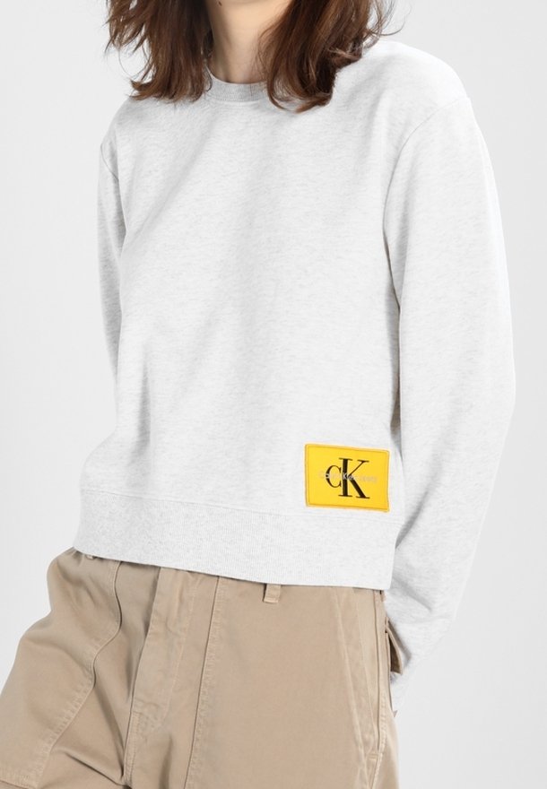 Sudadera de algodón gris claro con puños y dobladillo acanalados, que presenta una etiqueta rectangular amarilla prominente con un logotipo negro en el lado izquierdo.