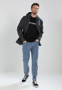 Donkergrijze outdoorjas met een capuchon over een zwart t-shirt met een witte logo, gecombineerd met lichtblauwe jeans en zwarte sneakers.