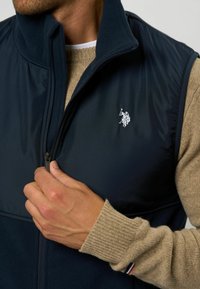 Marinblå zip-väst med fleecekrage, med en broderad logotyp på bröstet. Västen är lager på lager över en beige stickad tröja.