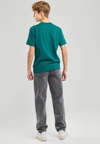 Camiseta de algodón teal con cuello redondo, mangas cortas y un corte holgado; combinada con jeans grises y zapatillas blancas.