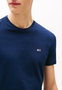 Tamsiai mėlynas medvilninis T-shirt su apvaliu kaklu ir trumpomis rankovėmis, prie krūtinės yra mažas siuvinėtasis logotipas raudonos, baltos ir mėlynos spalvos.