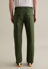 Groene katoenen broek met rechte snit, voorzien van twee achterzakken en een tailleband met riemlussen. Gedragen met tan en marineblauwe schoenen.