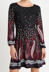 Robe noire avec des motifs floraux et paisley en rouge, blanc et violet. Manches longues, coupe évasée, texture douce et design à longueur genou.