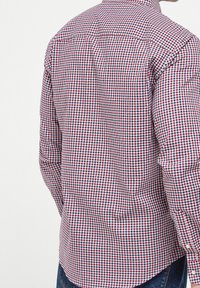 Camisa a cuadros con un patrón rojo, blanco y azul, con cuello abotonado y mangas largas. Fabricada con tela ligera.