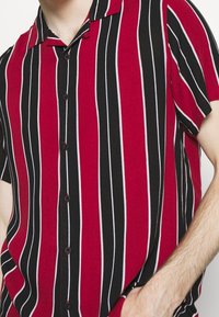 Mężczyzna w koszuli na krótki rękaw z pionowymi czerwonymi, czarnymi i białymi paskami, z ręką w kieszeni, na jednolitym tle.