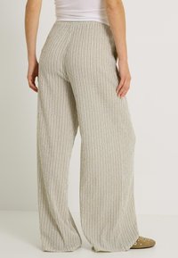 Mujer vistiendo pantalones de pierna ancha de rayas verticales en beige y blanco, y zapatos de slip-on marrones con eyelets metálicos, de pie frente a un fondo blanco.