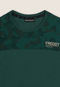 T-shirt vert à manches courtes avec un motif camo texturé sur la partie supérieure, arborant un logo et du texte beiges. Tissu doux avec un col rond.