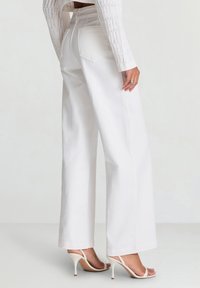 Jeans bianchi a gamba larga con vita alta, realizzati in tessuto robusto, con tasche posteriori. Abbinati a tacchi bianchi con cinturini.