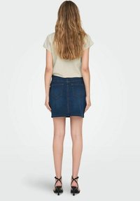 Džínová sukně v tmavě modré barvě s přiléhavým střihem, zadními kapsami a neupraveným lemem. Kombinováno se světlým tričkem s krátkými rukávy a černými lodičkami.