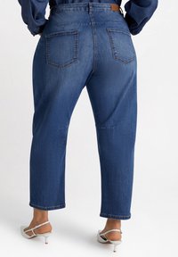 Jeans de tiro alto y pierna recta en denim azul oscuro, con dos bolsillos traseros. Combinados con sandalias blancas de tacón para un contraste.