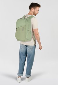 Zaino verde chiaro con un design aerodinamico, tasca frontale con zip e dettaglio del logo. Le spalline sono imbottite e viene portato su una camicia chiara.