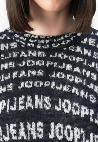 Strikkegenser i mørkeblå med hvitmønstret tekst. Rund hals og myk tekstur, med gjentakelse av "JEANS JOOP!" over hele stoffet.