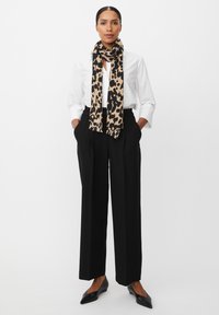Camicia bianca a bottoni, pantaloni neri a gamba larga e una sciarpa beige con stampa leopardata nera. Ballerine nere a punta, mani nelle tasche.