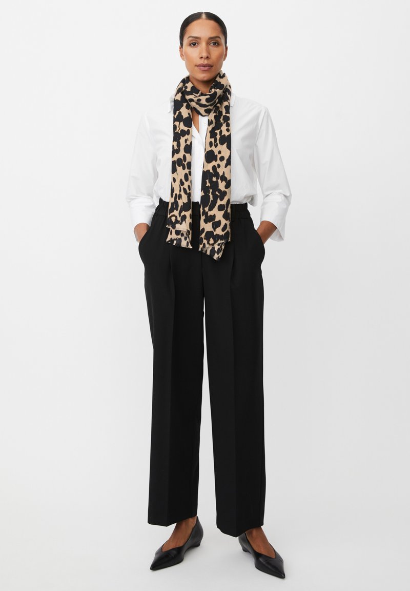Camicia bianca a bottoni, pantaloni neri a gamba larga e una sciarpa beige con stampa leopardata nera. Ballerine nere a punta, mani nelle tasche.