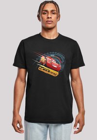 Melnā T-krekliņš ar Lightning McQueen grafiku sarkanā un dzeltenā krāsā, ar ziliem ģeometriskiem akcentiem un apaļu kakla izgriezumu.