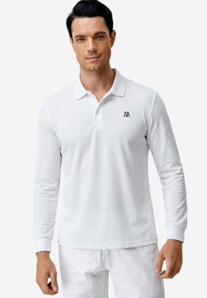 Mann trägt ein weißes langärmeliges Poloshirt mit kleinem schwarzem Logo auf der linken Brust und weiße Hose, steht vor einfarbigem Hintergrund.