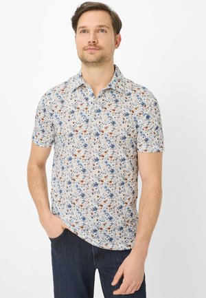 Man met kort haar die een wit overhemd met korte mouwen draagt met een blauw-bruine bloemenprint en donkerblauwe spijkerbroek, staand met één hand in de zak.