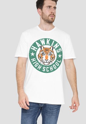 Mann trägt ein weißes T-Shirt mit einem grünen, kreisförmigen Hawkins High School-Logo mit einem Tigerkopf, kombiniert mit blauen Jeans.