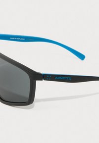 Occhiali da sole Arnette neri e blu con lenti rettangolari scure e la scritta "Fatti di bioplastica" stampata sull'asta blu.