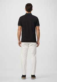 Muž s čiernym polokošilom a bielymi nohavicami, stojaci chrbtom k jednoliatej pozadí.