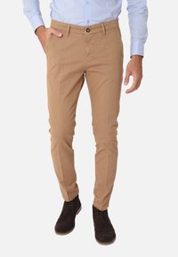 Chinos beige realizzati in cotone con un design slim fit, dotati di tasche laterali, chiusura con bottone e una texture sottile. Indossati con stivali scuri.