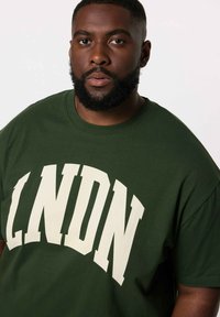 Grøn bomulds T-shirt med store hvide "LNDN" bogstaver. Korte ærmer, rund halsudskæring, afslappet pasform, glat tekstur.