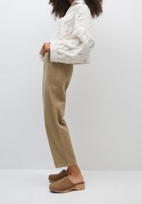 Pull en tricot blanc avec motif torsadé, pantalon en velours côtelé beige et sabots en bois marron. Vue de profil, montrant une coupe décontractée et la texture.