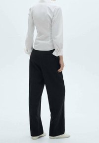 Chemise blanche à col avec manches longues légèrement bouffantes, associée à un pantalon noir ample et des chaussures de couleur claire. Fond uni.