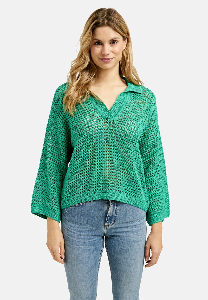 Grüner gestrickter Pullover mit lockerem, offenem Gewebe, Kragendesign und weiten Ärmeln. Kombiniert mit hellblauen Jeans, die einen lässigen Look zeigen.