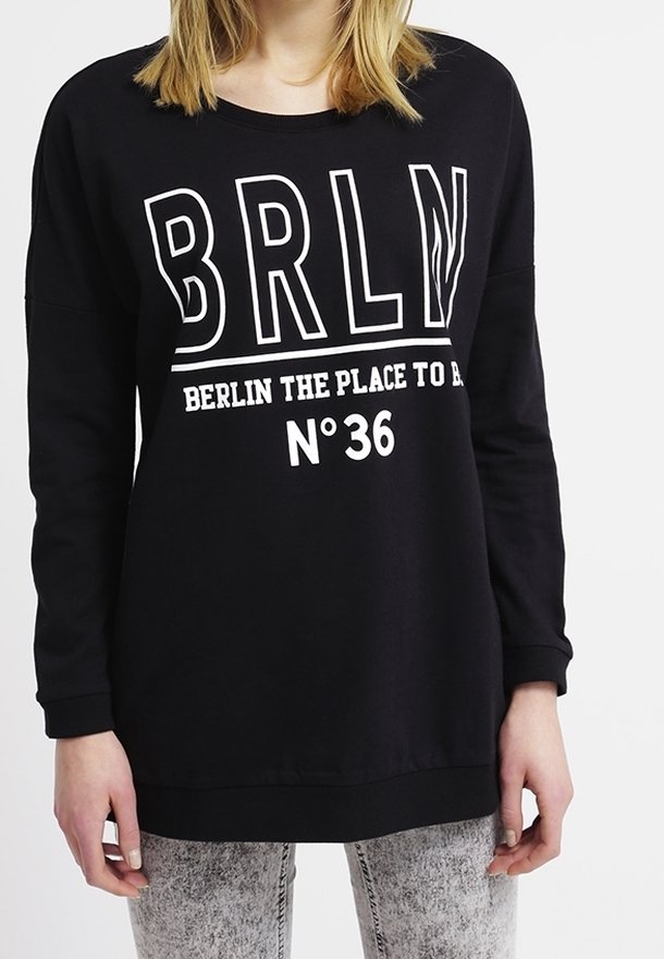 Chemise noire à manches longues avec texte blanc "BRLN" et "Berlin the place to be N°36" portée par une personne avec un jean clair.